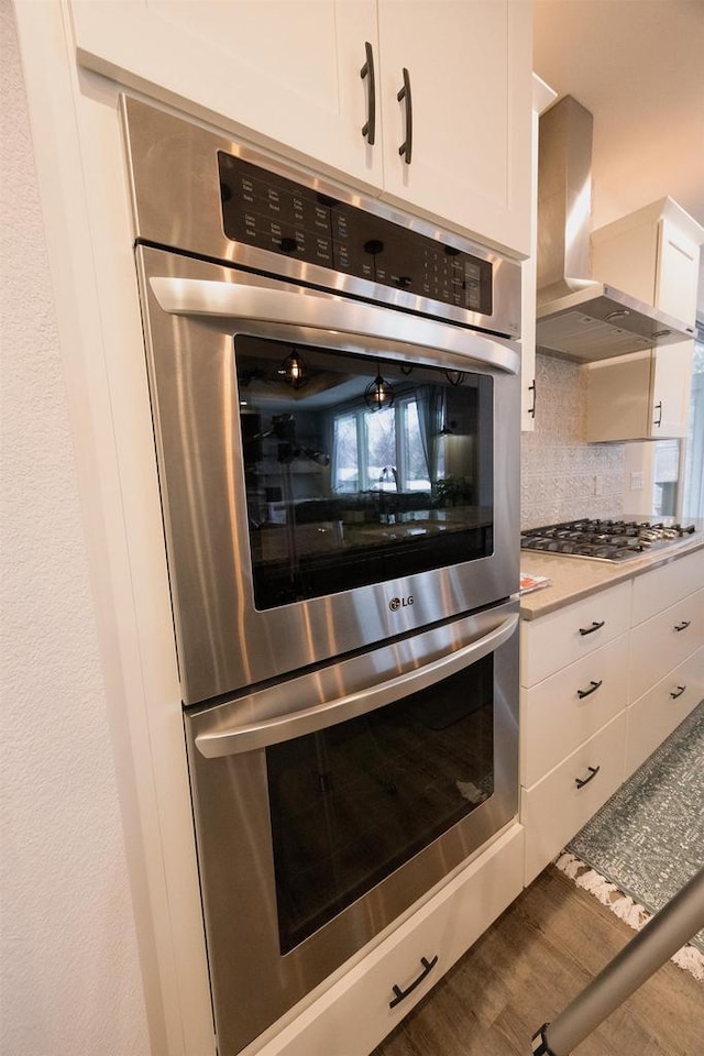 kitchen featuring stainless steel appliances, wood finished floors, white cabinetry, decorative backsplash, and wall chimney exhaust hood