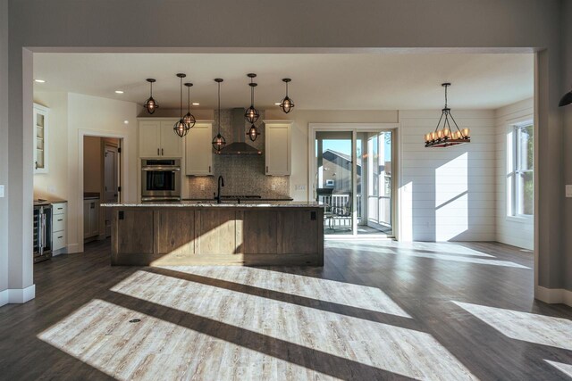 kitchen featuring oven, wall chimney exhaust hood, tasteful backsplash, an island with sink, and dark wood finished floors