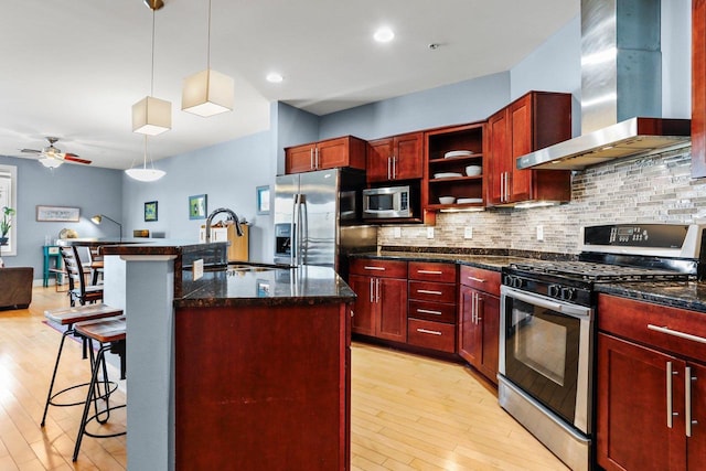 kitchen with appliances with stainless steel finishes, decorative light fixtures, wall chimney range hood, sink, and a center island with sink