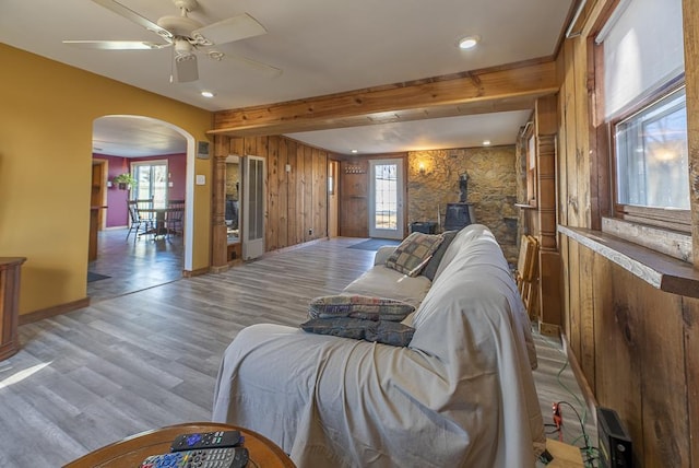 living room featuring wooden walls, arched walkways, beamed ceiling, a wood stove, and light wood-style floors