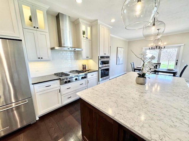 kitchen with backsplash, pendant lighting, appliances with stainless steel finishes, wall chimney exhaust hood, and dark stone counters