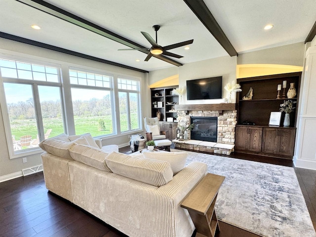 living room with a stone fireplace, ceiling fan, beamed ceiling, dark wood-type flooring, and built in shelves