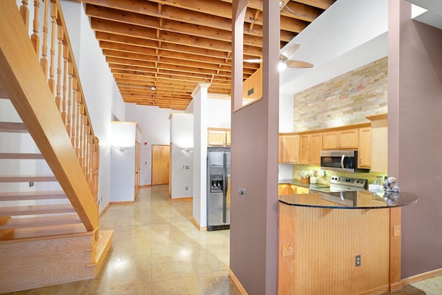 kitchen with light brown cabinetry, ceiling fan, a towering ceiling, dark stone countertops, and stainless steel appliances