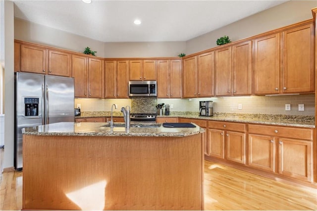 kitchen with appliances with stainless steel finishes, an island with sink, sink, light stone counters, and light wood-type flooring