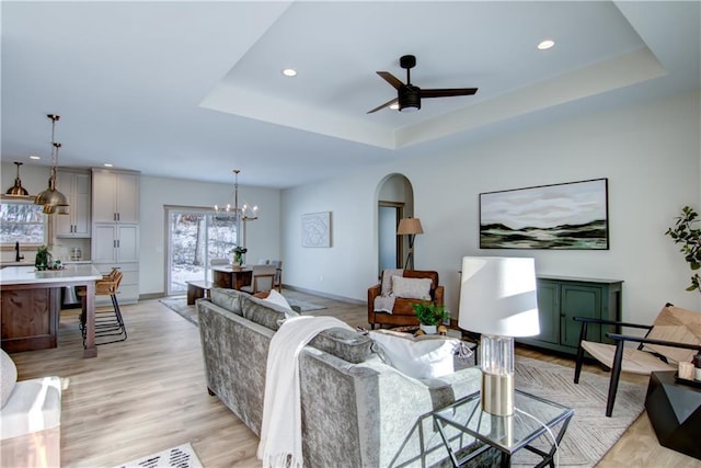 living room with light wood-type flooring, ceiling fan with notable chandelier, and a tray ceiling