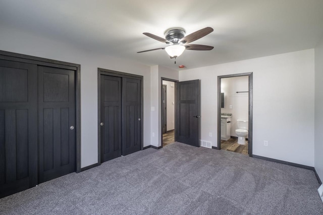 unfurnished bedroom with dark colored carpet, two closets, ensuite bath, and ceiling fan