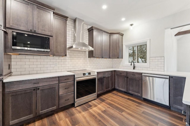 kitchen featuring appliances with stainless steel finishes, decorative backsplash, wall chimney range hood, sink, and dark hardwood / wood-style flooring