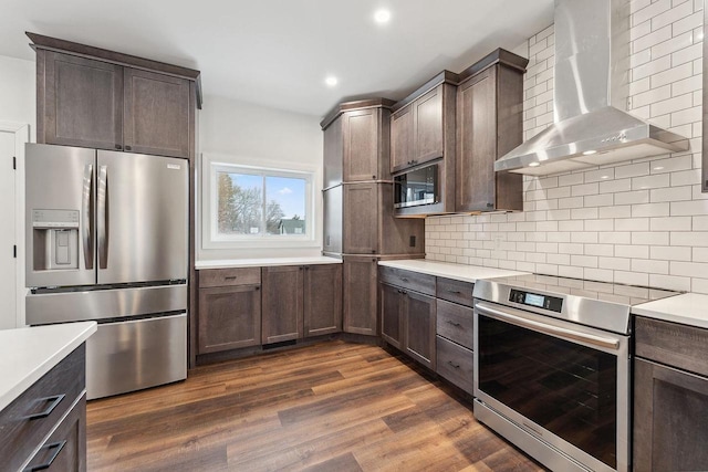 kitchen with dark brown cabinetry, wall chimney exhaust hood, stainless steel appliances, backsplash, and dark hardwood / wood-style floors