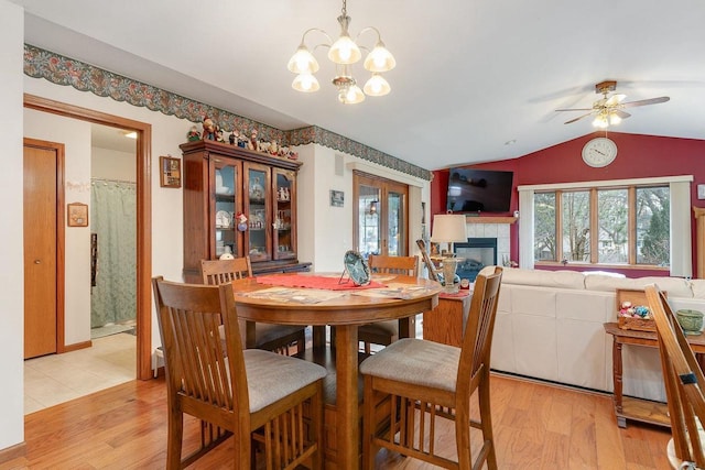 dining room with a fireplace, ceiling fan with notable chandelier, vaulted ceiling, and light wood-type flooring