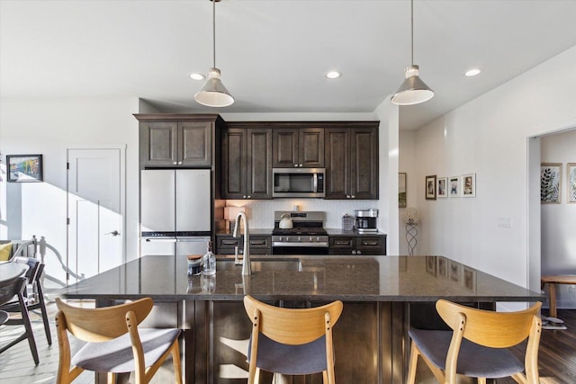 kitchen with dark brown cabinets, appliances with stainless steel finishes, a kitchen island with sink, and hanging light fixtures