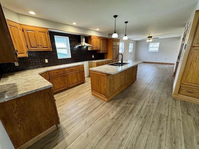 kitchen with sink, hanging light fixtures, light wood-type flooring, a kitchen island with sink, and wall chimney range hood