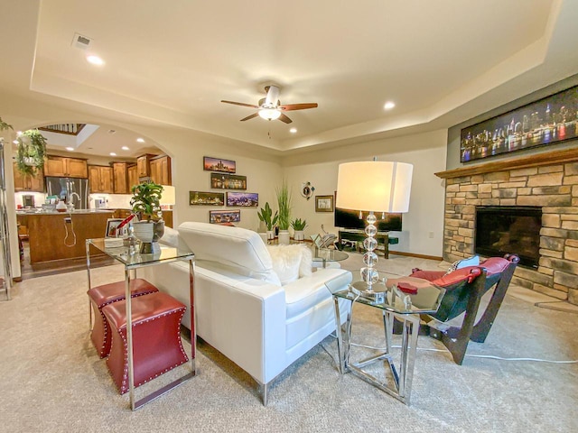 carpeted living room with ceiling fan, a tray ceiling, and a stone fireplace