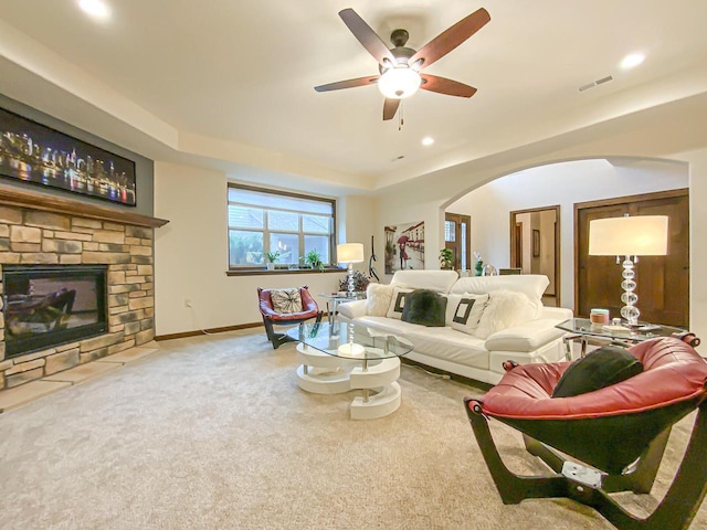 carpeted living room featuring a tray ceiling, a stone fireplace, and ceiling fan