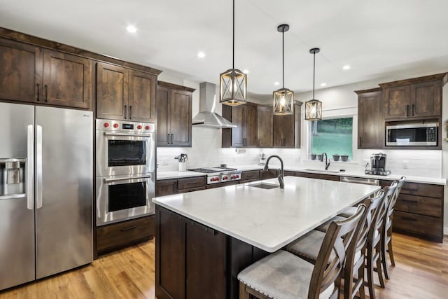 kitchen featuring pendant lighting, an island with sink, wall chimney exhaust hood, stainless steel appliances, and light hardwood / wood-style flooring