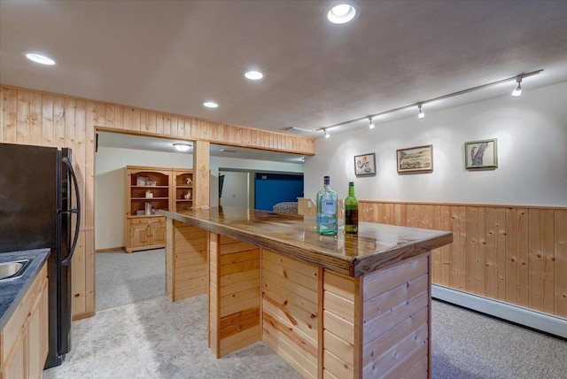 kitchen with light brown cabinetry, black refrigerator, a baseboard radiator, wooden walls, and light colored carpet