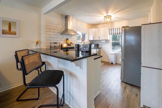 kitchen with tasteful backsplash, white cabinetry, a kitchen bar, stainless steel appliances, and wall chimney exhaust hood