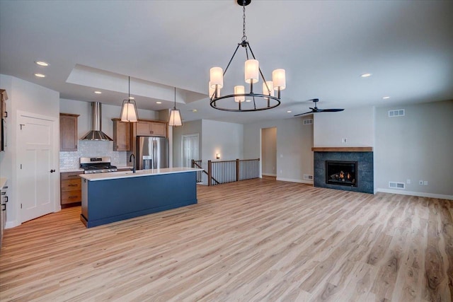 kitchen with a kitchen island with sink, decorative light fixtures, appliances with stainless steel finishes, wall chimney range hood, and light hardwood / wood-style floors