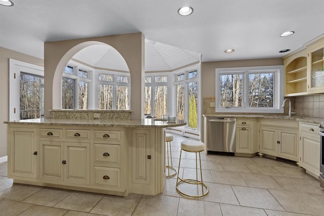 kitchen with a kitchen bar, dishwasher, cream cabinetry, light stone countertops, and backsplash