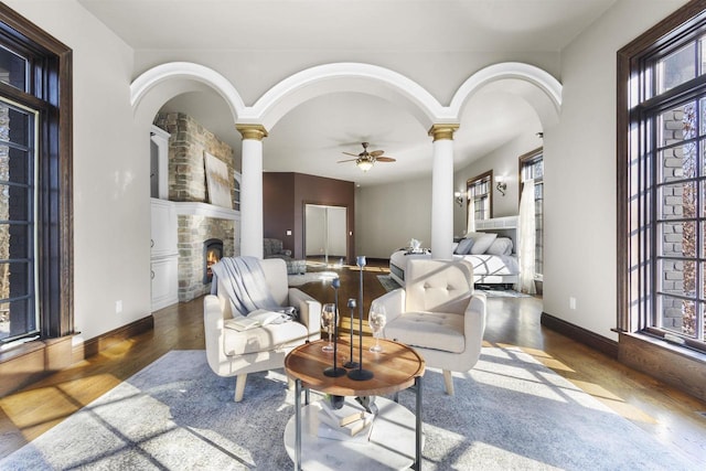 living room with ceiling fan, wood-type flooring, a stone fireplace, and ornate columns