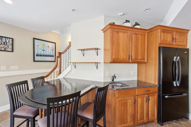 kitchen with brown cabinets, recessed lighting, freestanding refrigerator, a sink, and dark stone countertops