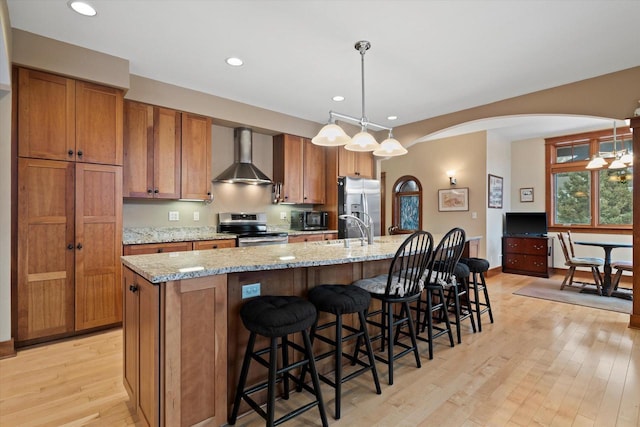 kitchen featuring wall chimney range hood, a kitchen island with sink, hanging light fixtures, stainless steel appliances, and a kitchen bar