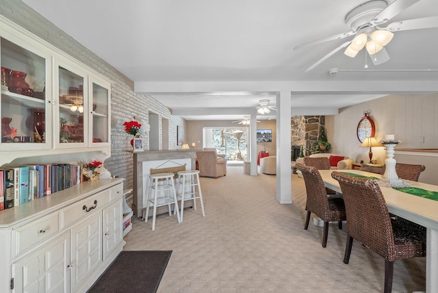 dining space with beam ceiling, a stone fireplace, light colored carpet, and ceiling fan