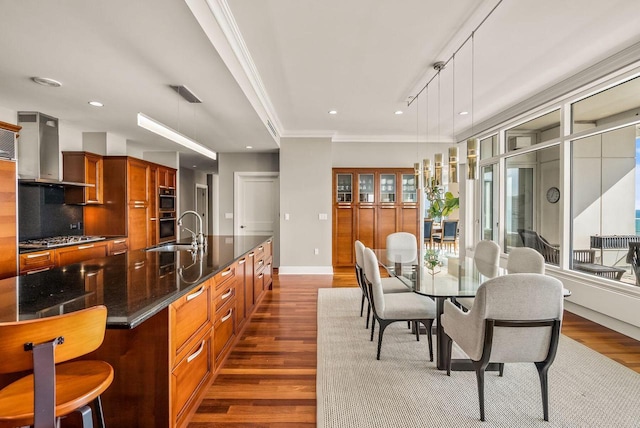 kitchen with wall chimney exhaust hood, sink, appliances with stainless steel finishes, an island with sink, and pendant lighting