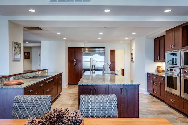 kitchen with built in appliances, sink, light stone counters, and an island with sink