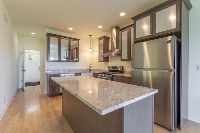 kitchen with wall chimney exhaust hood, hanging light fixtures, a kitchen island, stainless steel appliances, and light stone countertops