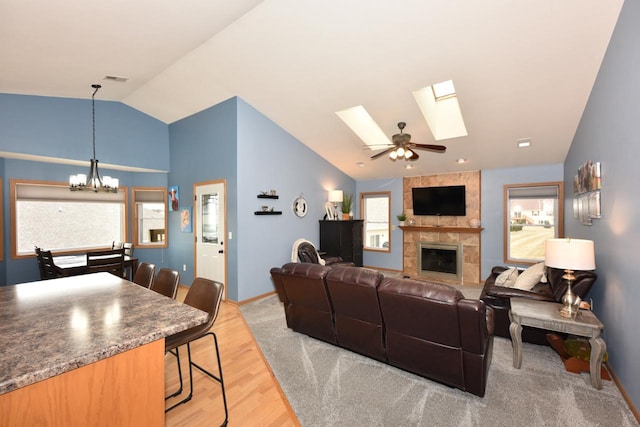 living room featuring a tile fireplace, lofted ceiling with skylight, ceiling fan with notable chandelier, and light wood-type flooring