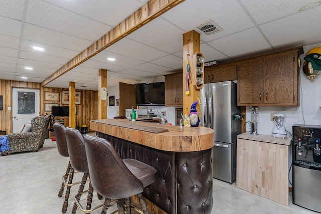 kitchen with a breakfast bar, a drop ceiling, stainless steel fridge, and wood walls