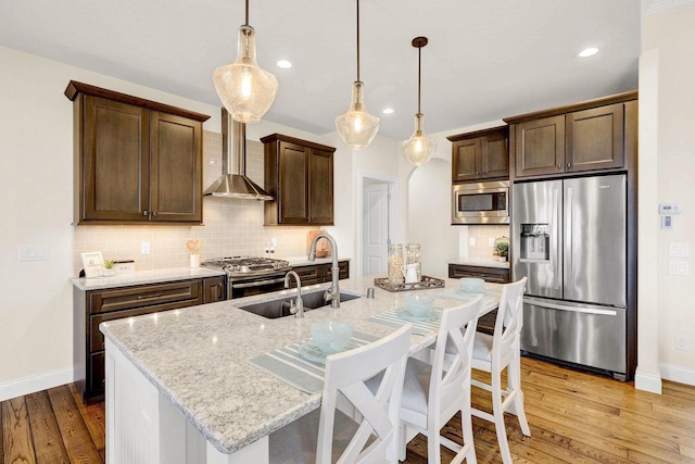 kitchen with wall chimney range hood, sink, hanging light fixtures, stainless steel appliances, and an island with sink