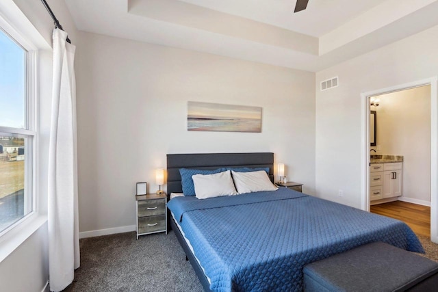 bedroom with a raised ceiling, dark colored carpet, connected bathroom, and ceiling fan