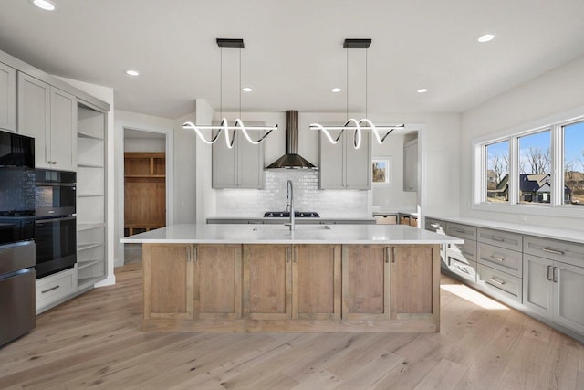 kitchen with light hardwood / wood-style flooring, a kitchen island with sink, pendant lighting, and wall chimney exhaust hood