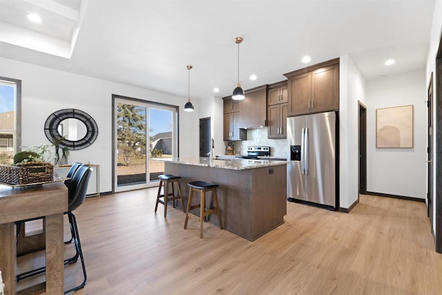 kitchen with light stone counters, decorative light fixtures, light wood-type flooring, appliances with stainless steel finishes, and a kitchen island with sink