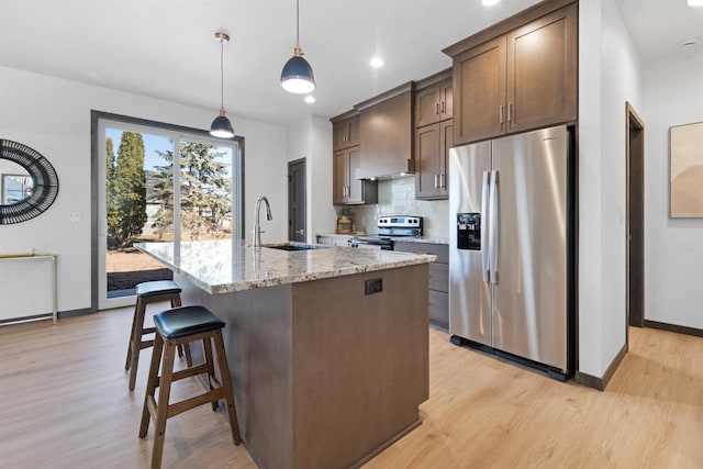 kitchen with pendant lighting, sink, stainless steel appliances, light stone counters, and a center island with sink