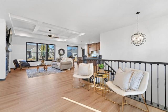 living room with coffered ceiling, a stone fireplace, light hardwood / wood-style floors, beam ceiling, and ceiling fan with notable chandelier