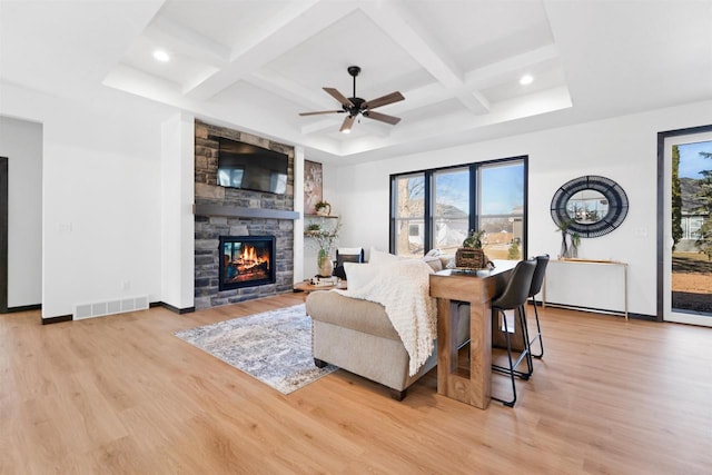 living room with ceiling fan, coffered ceiling, light hardwood / wood-style floors, a stone fireplace, and beamed ceiling