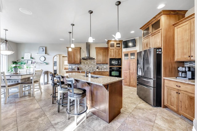 kitchen with sink, decorative light fixtures, black appliances, a center island with sink, and wall chimney range hood