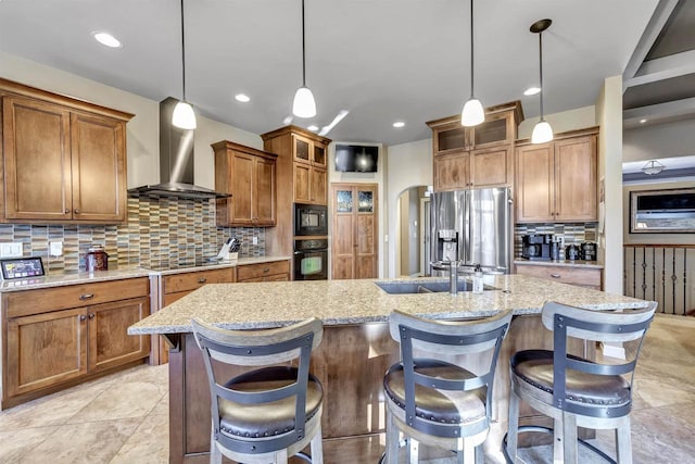 kitchen with an island with sink, a kitchen breakfast bar, hanging light fixtures, black appliances, and wall chimney exhaust hood