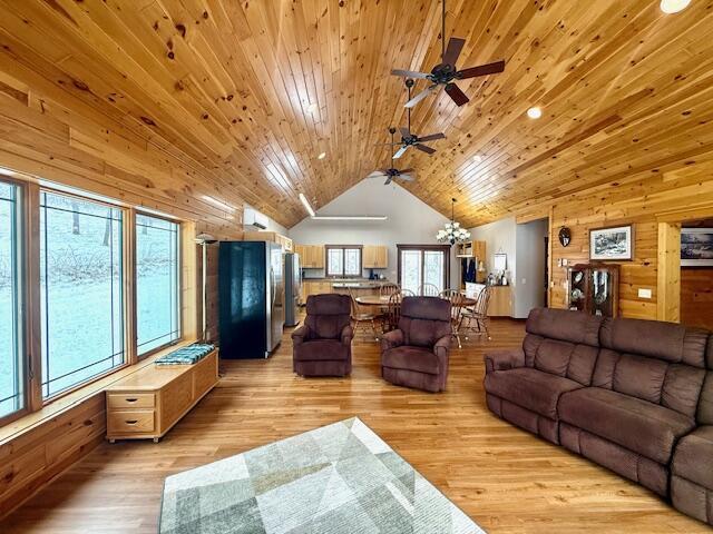 living room with ceiling fan with notable chandelier, high vaulted ceiling, wooden walls, wood ceiling, and light hardwood / wood-style flooring