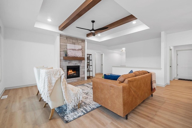 living room with ceiling fan, a stone fireplace, a raised ceiling, beamed ceiling, and light wood-type flooring