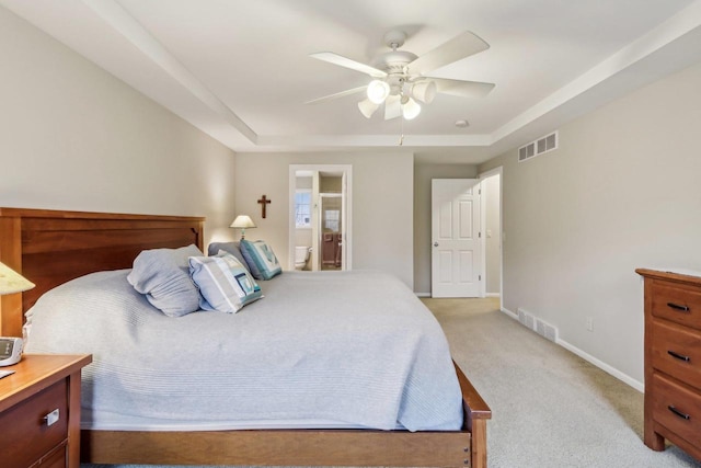 bedroom with a raised ceiling, light colored carpet, ceiling fan, and ensuite bath