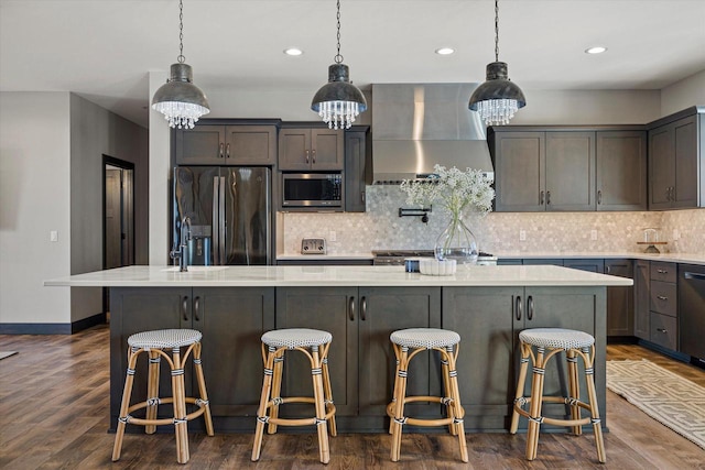 kitchen with an island with sink, hanging light fixtures, appliances with stainless steel finishes, wall chimney range hood, and a kitchen breakfast bar