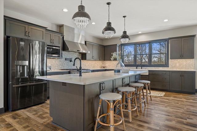 kitchen featuring a center island with sink, sink, decorative light fixtures, stainless steel appliances, and wall chimney exhaust hood