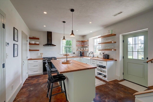 kitchen with wall chimney range hood, butcher block counters, a center island, white cabinets, and stainless steel range with gas cooktop