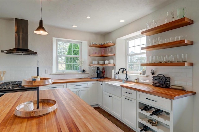 kitchen with hanging light fixtures, white cabinetry, butcher block counters, and wall chimney range hood