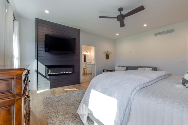 bedroom featuring connected bathroom, a large fireplace, ceiling fan, and light wood-type flooring