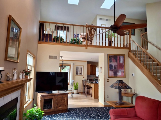 living room with light hardwood / wood-style flooring, a tile fireplace, ceiling fan, a skylight, and a high ceiling