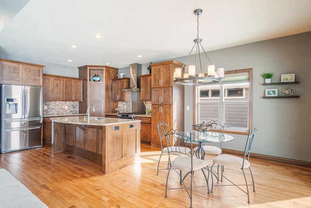 kitchen with pendant lighting, a kitchen island with sink, light stone counters, stainless steel fridge with ice dispenser, and wall chimney range hood